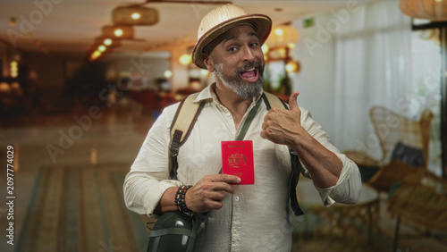 Принти на полотні Man with backpack and pith helmet holding passport and pointing thumb to it in a hotel building lobby; adventure excitement