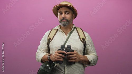 Foto Man holding binoculars and backpack, wearing pith helmet and canvas shirt, winking and facing camera in a pink studio; curiosity exploration adventure