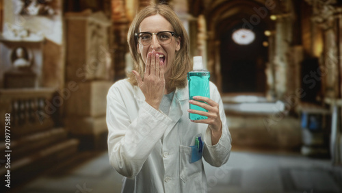 Canvas Print Woman doctor in white coat holding a turquoise mouthwash bottle while covering mouth with hand and smiling in an ornate building corridor; playful hygiene demo
