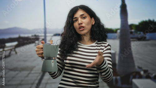 Εκτύπωση καμβά Young woman holding a moka pot with one hand and speaking while gesturing with the other on a seaside street; casual