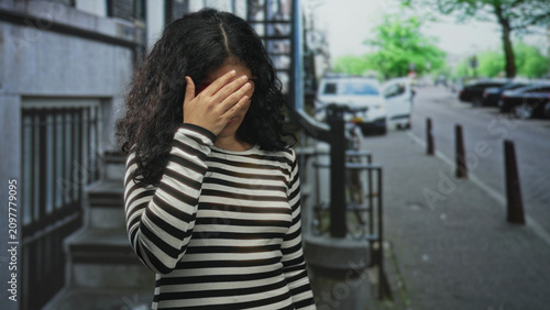 Foto Woman with hands covering her face on a city street beside a stair entrance and parked cars; embarrassment privacy vulnerability