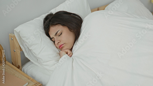 Young hispanic woman holding white duvet with one hand, eyes closed and face resting on pillow while settling into sleep on a bed in a bedroom; calm.