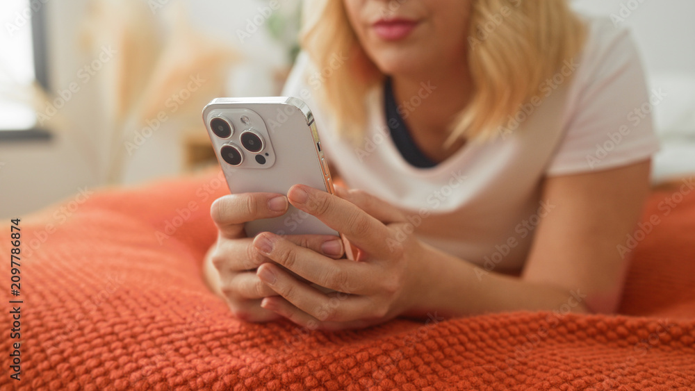 Fototapeta premium Young blonde woman holding smartphone with both hands, thumbs tapping screen on orange bedspread in bedroom; relaxation.