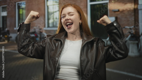Fotografie Redhead woman wearing leather jacket and white crop top raises clenched fists in victory on street; joy triumph celebration