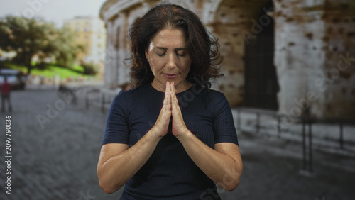 Tableau sur toile Woman with hands pressed in prayer, eyes closed, standing before an ancient building and cobblestone plaza in a tourist square; serenity faith