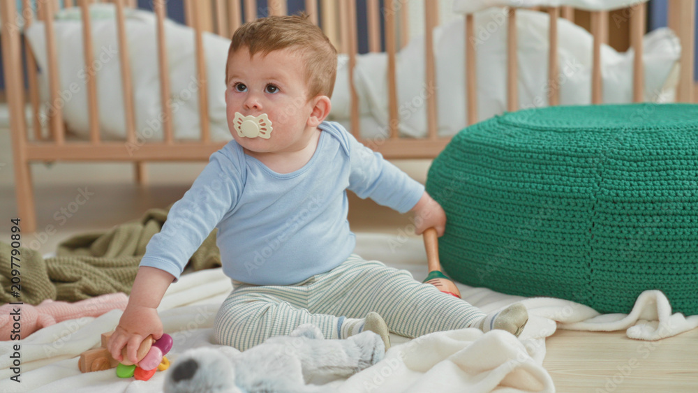Fototapeta premium Toddler boy holds colorful ring toy with pacifier in mouth while sitting on blanket near crib in nursery; curiosity comfort.
