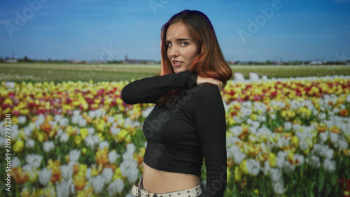 Fotografie Woman with red hair holding her shoulder and wincing in a tulip field, cropped midriff and studded belt visible; discomfort concern