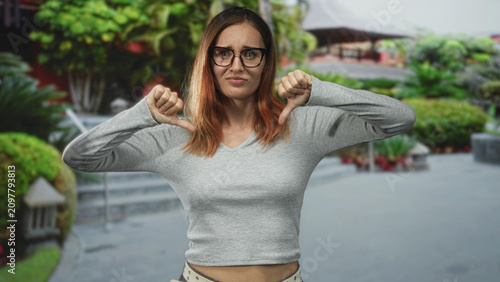 Fotografi Woman with glasses showing thumbs down gesture on a street with steps and greenery; disapproval rejection