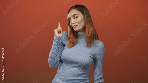 Fotografía Woman points index finger upward near temple with visible hand and freckles, wearing blue sweater against red studio wall; playful confidence