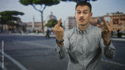 Fotografie Man with moustache and checked shirt shows both middle fingers in street square with stone pavement and umbrella pine trees, confrontational gaze; anger defiance