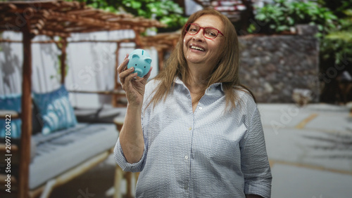 Obraz na plátne Senior woman holding blue piggy bank in right hand and smiling in a building patio with pergola and stone wall; saving retirement contentment