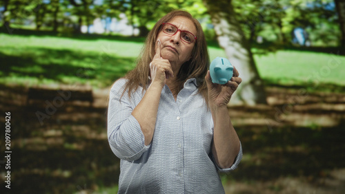 Obraz na plátne Woman holds blue piggy bank and touches chin while considering savings, standing in a forest park by terraced stone steps; retirement worry