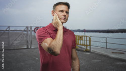 Принти на полотні Handsome hispanic man with hand on cheek wincing near pier railing and yellow barrier by the water; toothache pain