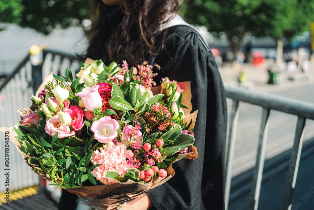 Fototapeta premium A side profile of a university graduate wearing a black academic gown and holding a vibrant flower bouquet filled with pink roses, carnations, and lush green foliage, positioned next to a metal bridge