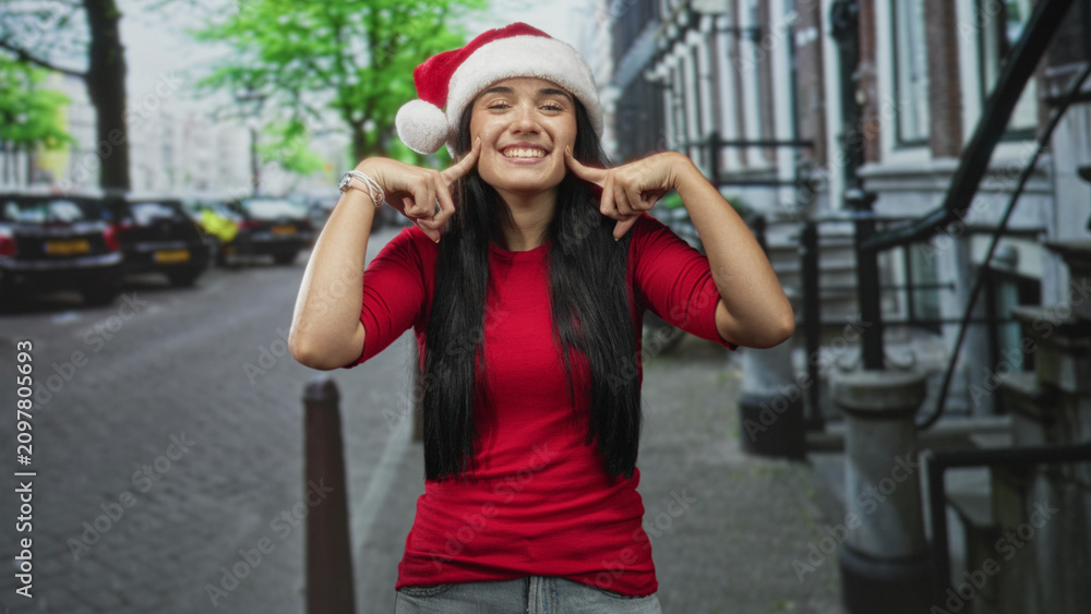 Fototapeta premium Young woman in santa hat presses cheeks with index fingers, smiling broadly on a city street; holiday cheer joyful.