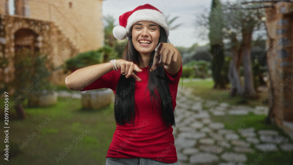 Fototapeta premium Woman pointing finger to camera with hand and smiling while wearing santa hat and red shirt in a historic stone courtyard with arched building and path; holiday joy.