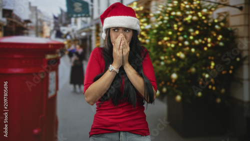 Фотография Woman in santa hat crosses hands over chest and shivers on a festive street by a christmas tree and red postbox; anxiety solitude