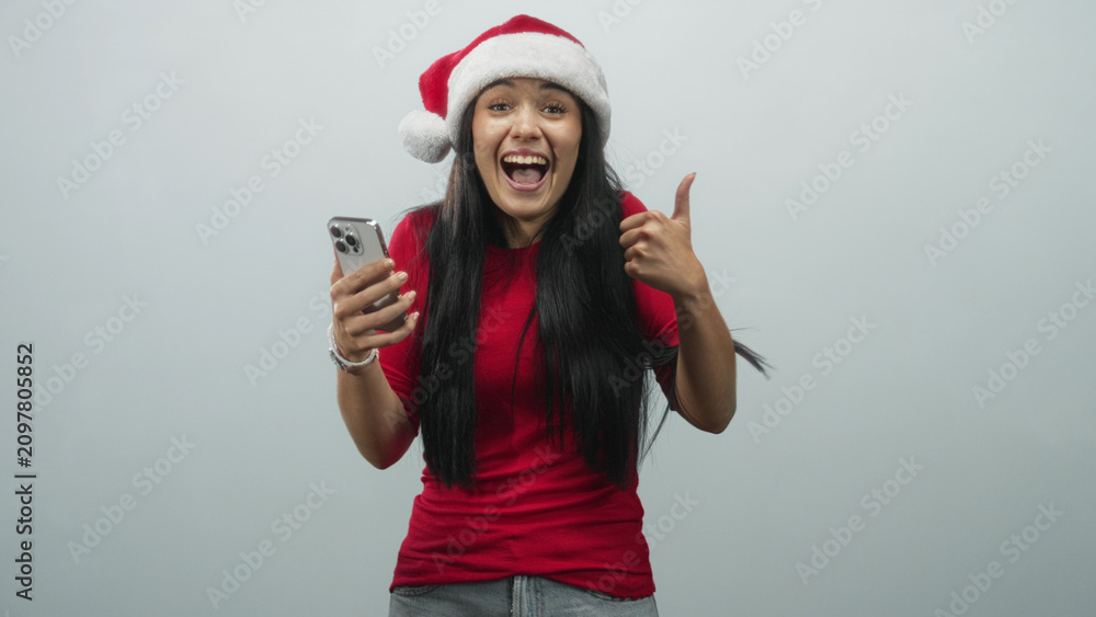 Fototapeta premium Young woman in santa hat holding smartphone and showing thumbs up in studio, wearing red shirt; joyful holiday celebration.