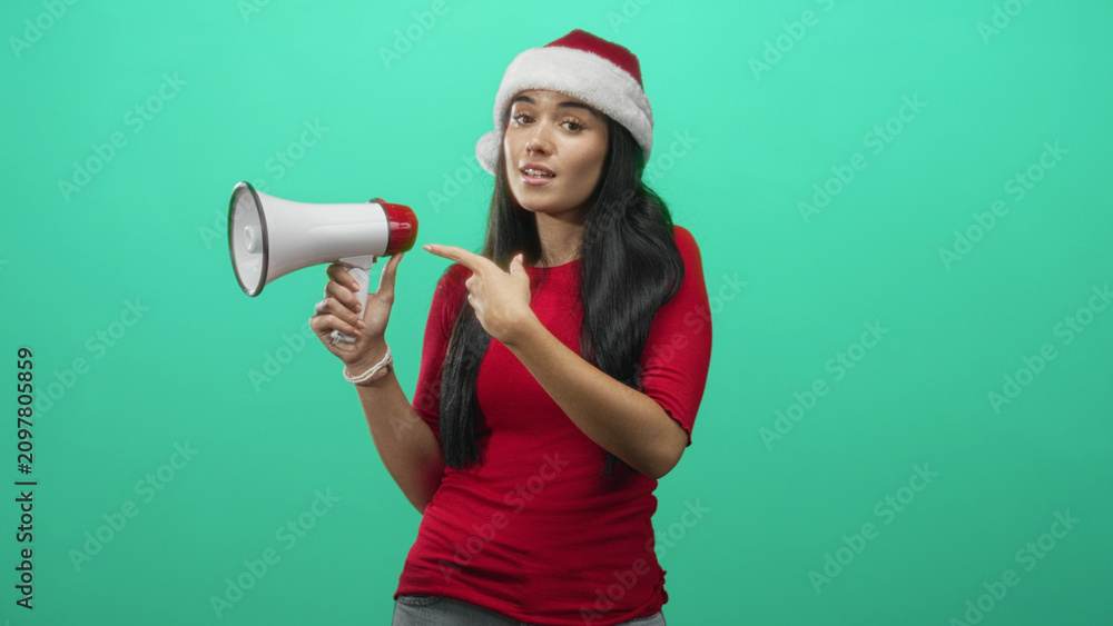 Fototapeta premium Woman in santa hat pointing finger at megaphone in studio, red top and exposed hand gesture for announcement; joyful holiday spirit.