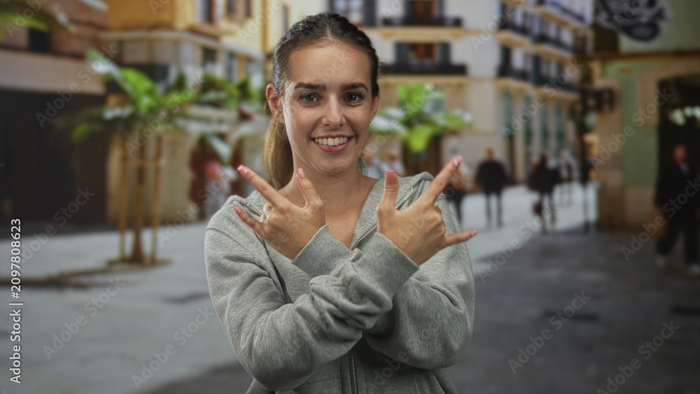 Fototapeta premium Young hispanic woman in gray hoodie smiling and making rock horns with both hands on a bustling city street; playful joy.