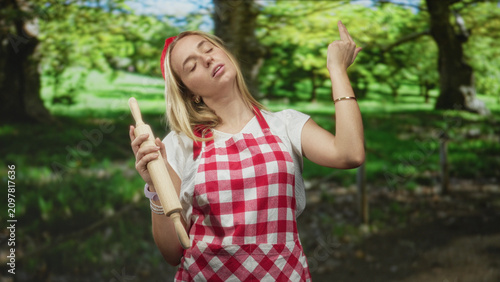 Konstfotografi Woman chef, blonde, wearing red gingham apron and headband, holding a wooden rolling pin, tilting head and flicking fingers in a forest setting; boredom