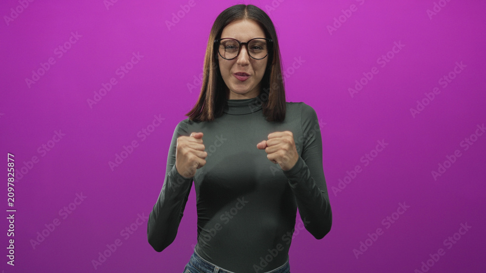 Fototapeta premium Young woman with glasses clenches fists and smiles in studio against purple backdrop; joy celebration.