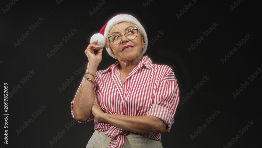 Fototapeta premium Woman in red striped shirt wearing glasses touches santa hat pompom to her ear while arms are crossed in studio; nostalgic holiday reflection.
