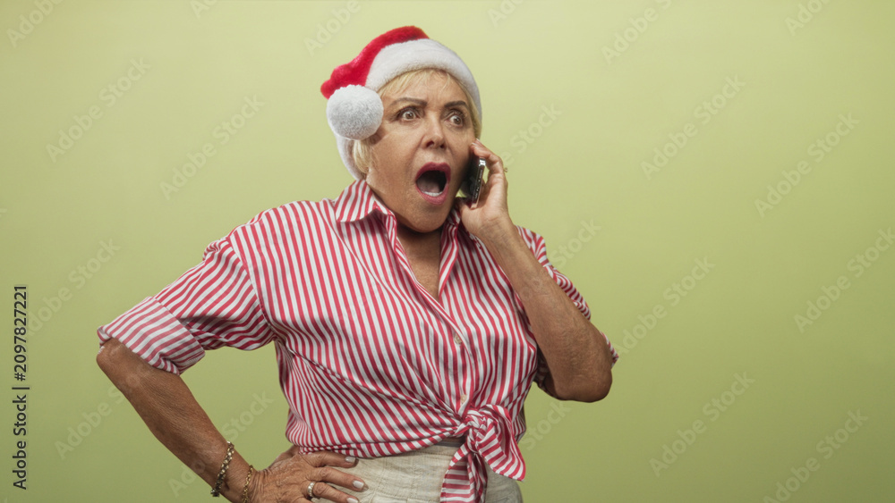 Fototapeta premium Woman, senior, wearing red santa hat and red striped shirt, holding smartphone to ear with hand on hip in studio; holiday surprise.