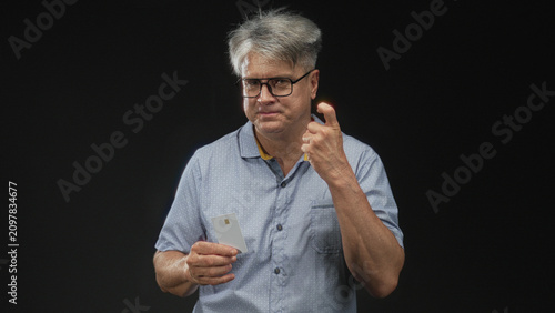 Fotografie Man with grey hair and glasses holding creditcard and pointing finger while smiling in studio; confidence convenient payment acceptance