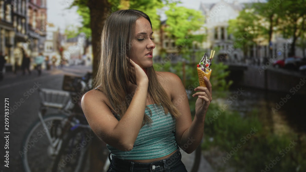 Fototapeta premium Woman holding an ice cream cone, hand to neck while inspecting dessert on a busy cobblestone street beside a canal with bicycles and trees; hesitation.