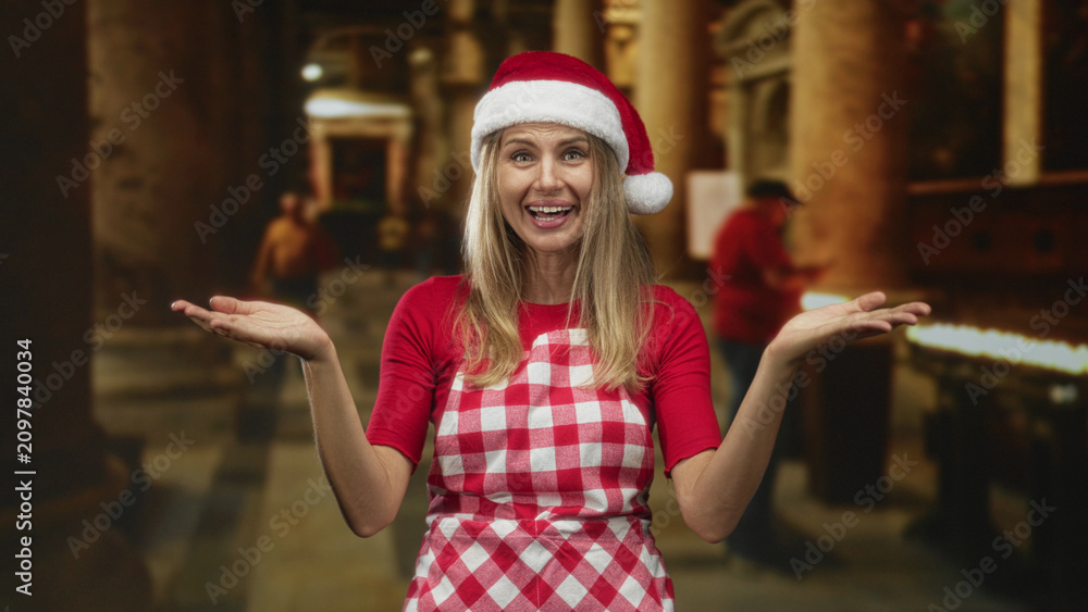 Fototapeta premium Woman wearing santa hat and red checkered apron shows open palms and smiles while shrugging inside a dim building aisle with candles and pillars; holiday cheer.