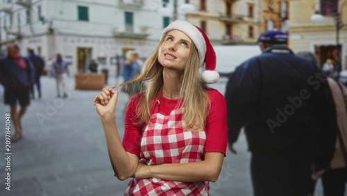 Quadro su tela Woman in santa hat and checked apron twirls hair with hand, arms crossed, standing on a street plaza; thoughtful holiday nostalgia