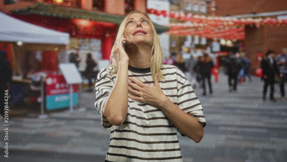 Fototapeta premium Woman on street holds phone to ear and clutches chest as if heartattack near outdoor market stalls and lanterns; urgent help distress.