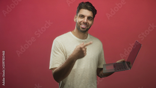 Cuadro en lienzo Smiling bearded man wearing striped tee holding laptop points finger to screen in studio; helpful online tutorial