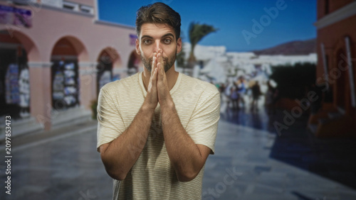 Foto Hispanic man with beard, hands clasped near mouth pleading on a sunny street lined with arched shops; appeal hope