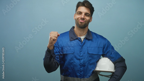 Fotografie Man construction worker in blue overalls holds white hardhat and makes fist pump gesture in studio; confidence achievement