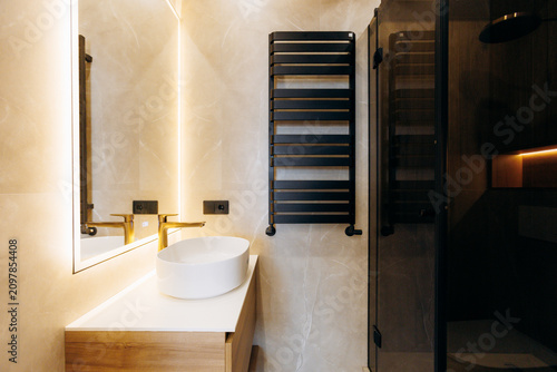 Modern bathroom vanity with round countertop sink, backlit mirror, warm ambient lighting, black heated towel rack, wooden cabinet, marble details, and compact minimal interior design