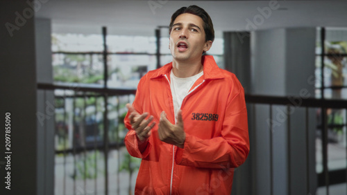 Obraz na plátne Man prisoner in orange jumpsuit clasping hands and looking upward beside an interior railing in a building, id number visible; remorse reflection penance