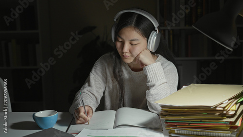 фотографія Woman writing in a notebook with headphones at a desk, pile of folders and mug under a lamp in a building interior; concentration quiet study