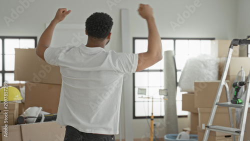 Tableau sur toile Man raising arms beside ladder, stacked moving boxes and cordless drill in building; moving day joy