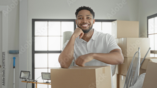 Foto Smiling man holds a cardboard box, rests arm on it and raises fist beside stacked boxes and floodlight in building; moving day confidence