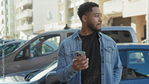 Canvastavla Man holding smartphone in right hand while glancing sideways near parked cars by apartment buildings on a street; thoughtful urban pause