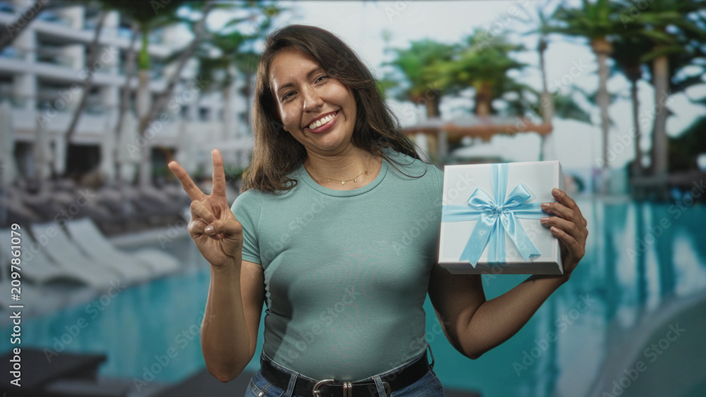 Fototapeta premium Woman in teal shirt holding wrapped gift box by pool makes peace sign and smiles with casual pose; celebration joy.