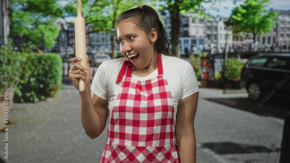 Fototapeta premium Woman cook smiling with eyes closed holds rolling pin upright on city street wearing red checkered apron and white tee; culinary joy.