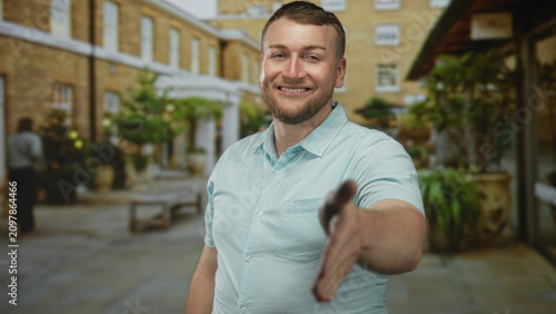 Cuadro en lienzo Man extending right hand for handshake toward camera in stone building courtyard, smiling and leaning forward as if meeting someone; friendly greeting