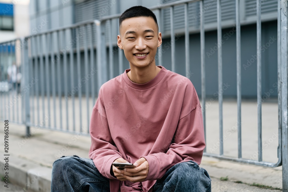 Obraz premium Young man seated by metal railing holding smartphone for casual portrait banner and social media background in urban public space