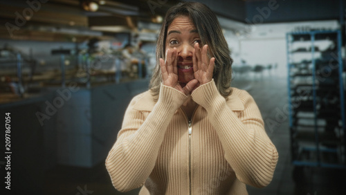 Fotografie Woman cupping hands to mouth shouting in bakery kitchen wearing beige zipper sweater and facing camera; urgency