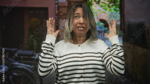 Foto Middle-aged hispanic woman grimacing with hands raised, mouth open in striped sweater on narrow cobblestone street with bicycle and building visible; stress