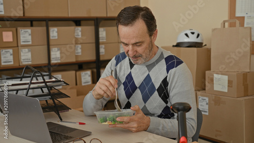 Konstfotografi Man holds fork eating salad from plastic container at desk with laptop and stacked cardboard boxes in storage building, hand visible; small business lunch calm