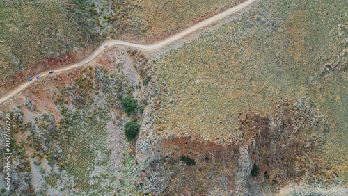 Top down aerial view of tourist cars driving on winding dirt road along steep mountain rocky slopes of Greece Mediterranean region in summer